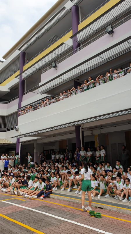 Students in green and white uniforms gather on a school courtyard and multi-level balconies during an outdoor event.